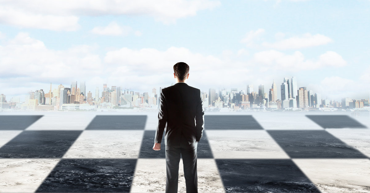 Strategic planning concept with businessman standing on chessboard and looking at city on sky background with clouds